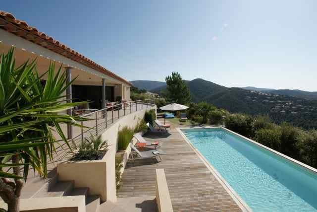 An outdoor area features a long heated swimming pool surrounded by a wooden deck. Lounge chairs are positioned for relaxation, with a shaded umbrella nearby. The villa’s expansive terrace opens towards panoramic views of the hills and mountains in the background.