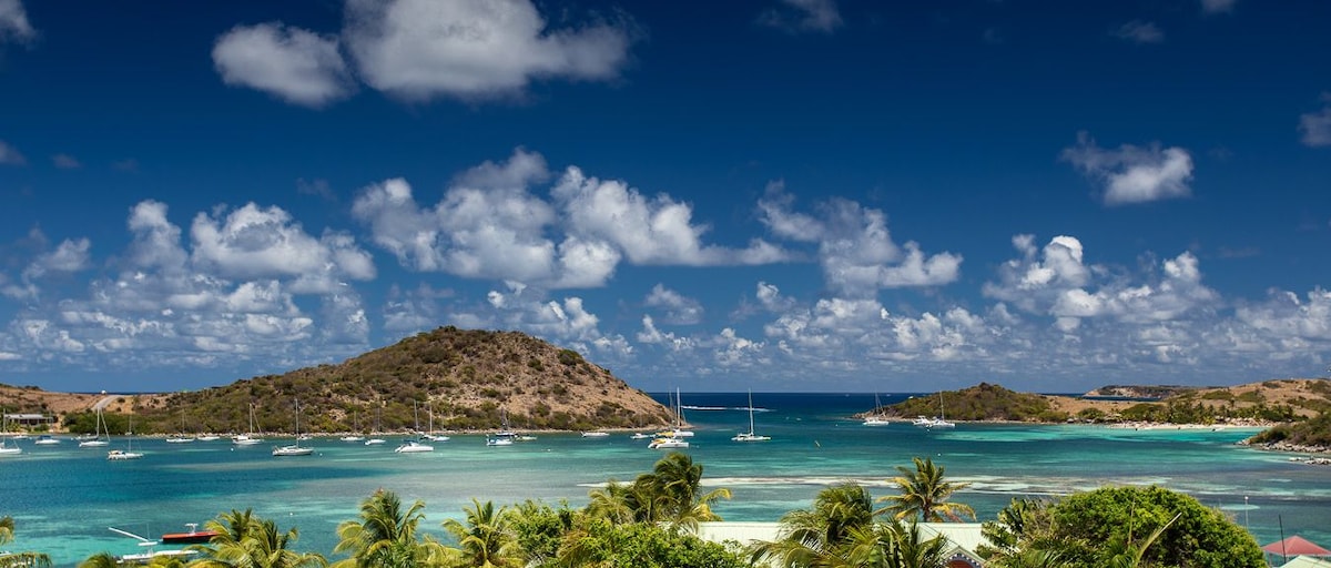 A panoramic view showcases the serene Bay of Cul de Sac, surrounded by lush green hills and dotted with sailboats. The vibrant turquoise waters reflect the blue sky, accented by white clouds, creating a tranquil coastal atmosphere.