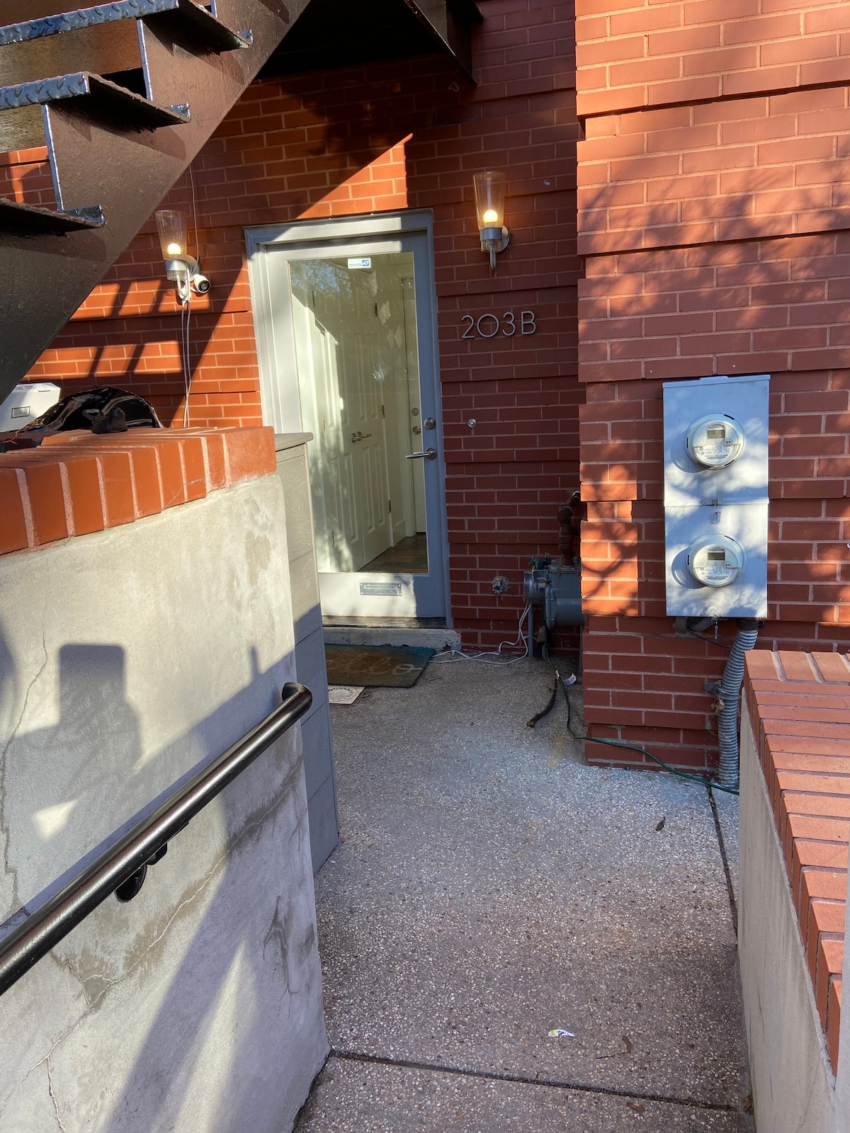 A pathway leads to a modern entry door marked '203B', framed by sleek light fixtures on a warm brick exterior. Nearby, utility meters are housed against the wall, and a concrete walkway extends toward the entrance, bordered by a railing and steps.