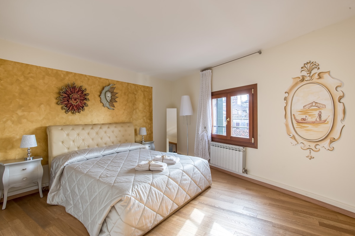 A spacious bedroom features a large, upholstered bed with a light-colored quilt. Decorative wall art and a window with sheer curtains are present, allowing natural light to illuminate the room. Light wooden flooring complements the warm tones of the walls.