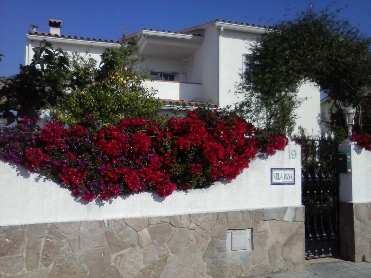 The entrance to the property is marked by a low stone wall adorned with vibrant flowering plants, dominated by shades of red and purple. A white house is visible in the background, characterized by its simple architecture and blue sky overhead.