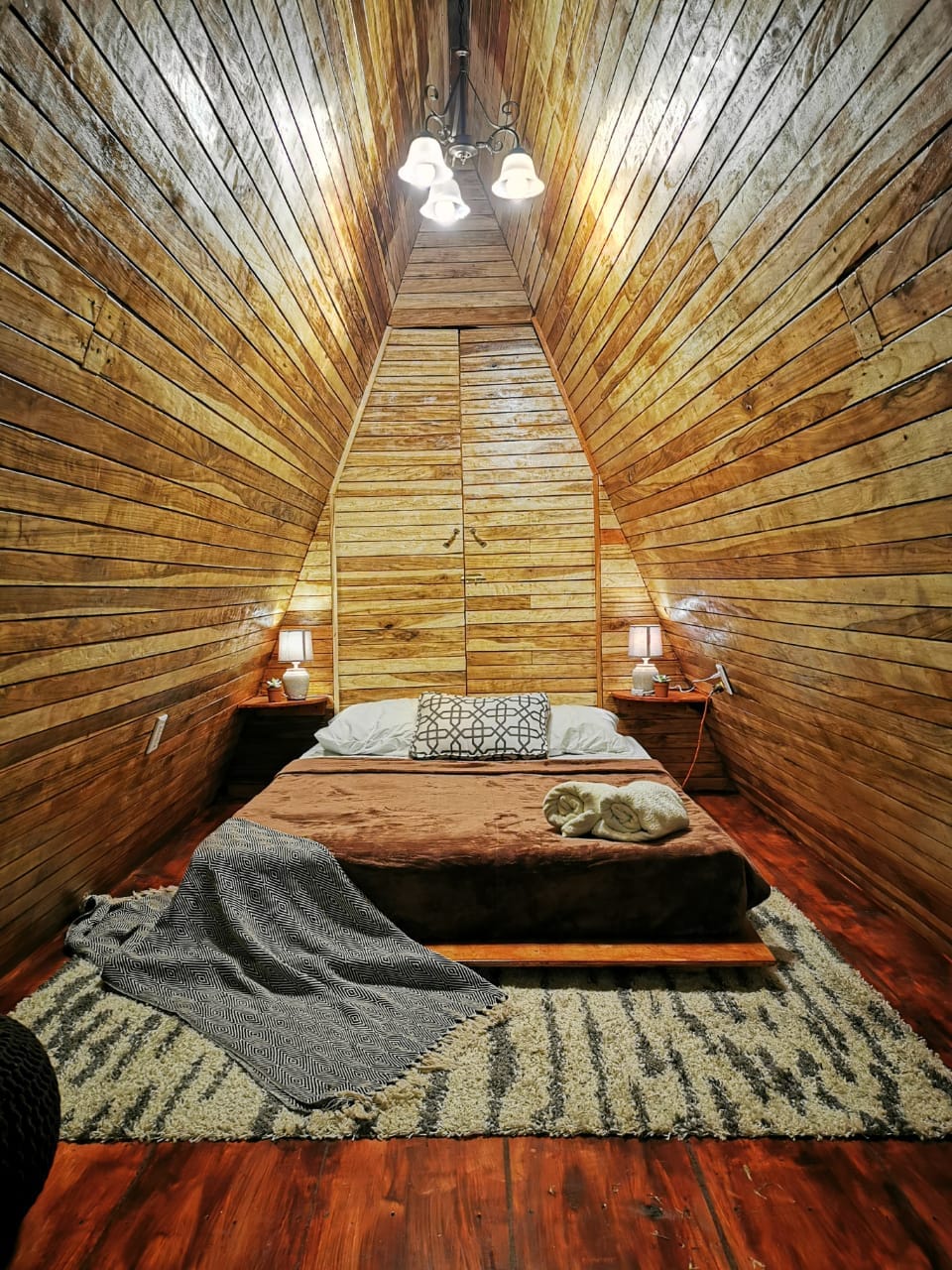 An intimate sleeping area is presented within a uniquely designed wooden structure. A spacious bed is topped with a neutral blanket and accompanied by a small side table. Soft lighting from wall lamps complements the warm wooden walls, enhancing the cozy ambiance.