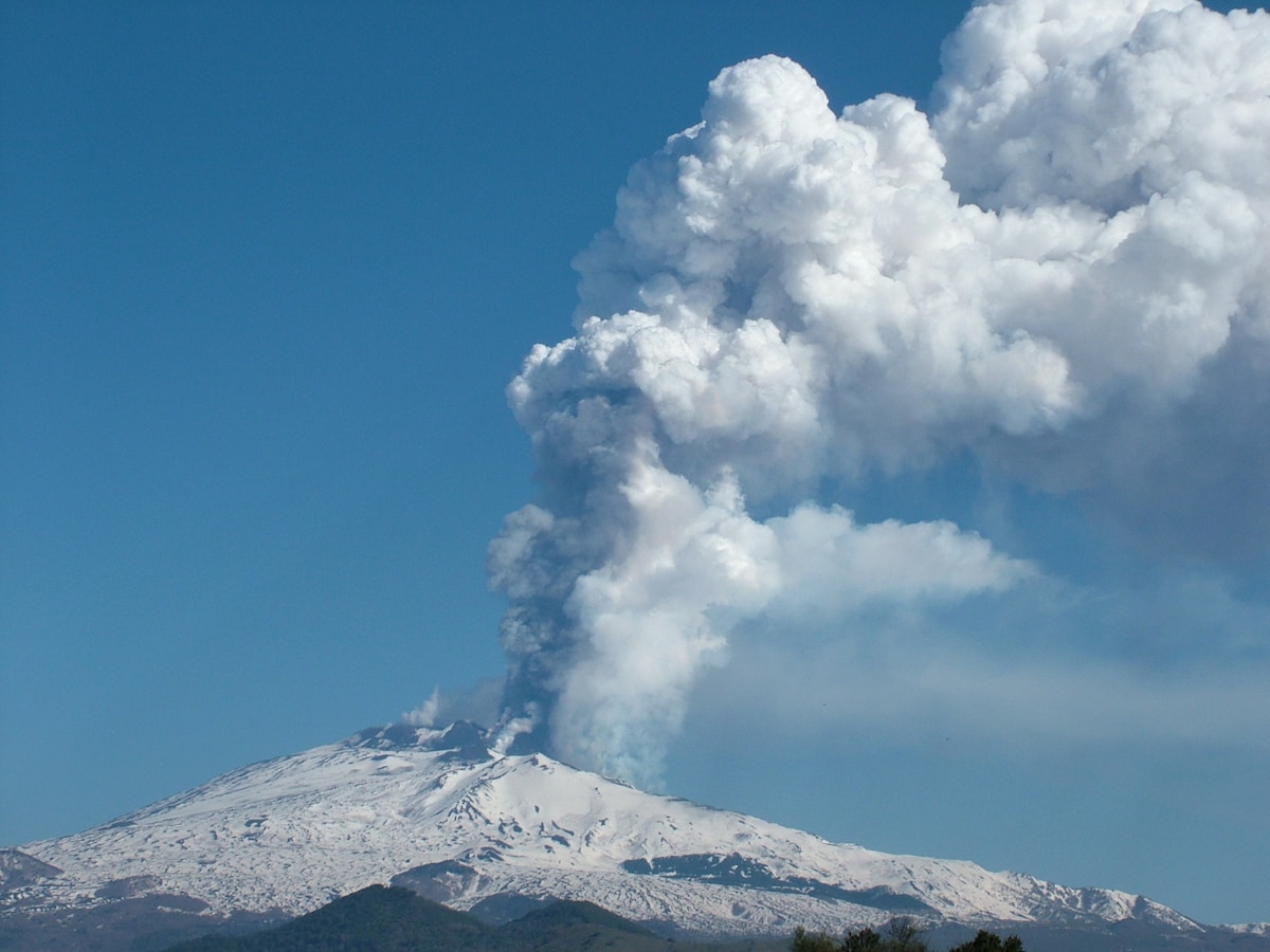 Mount Etna is depicted, with a snow-capped peak and a prominent plume of smoke rising from its summit. The sky is clear blue, contrasting with the white snow and gray ash, showcasing the natural beauty and volcanic activity of this UNESCO heritage site.