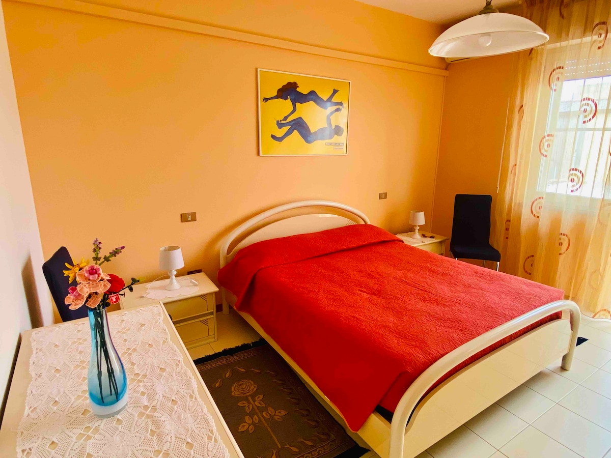A bright bedroom features a spacious double bed covered with a red blanket. Two bedside tables with lamps are positioned on either side of the bed. Soft, sheer curtains frame the window, allowing natural light to brighten the room. A decorative vase with flowers adds a touch of color.