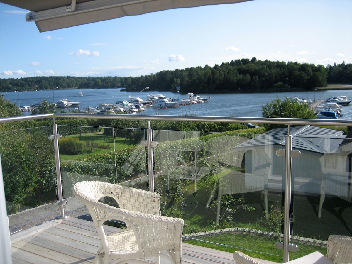 A balcony view showcases the serene water and surrounding greenery. Various boats can be seen docked in the distance, while comfortable white chairs invite relaxation. The scene is enhanced by clear skies and distant tree lines, creating a peaceful atmosphere.