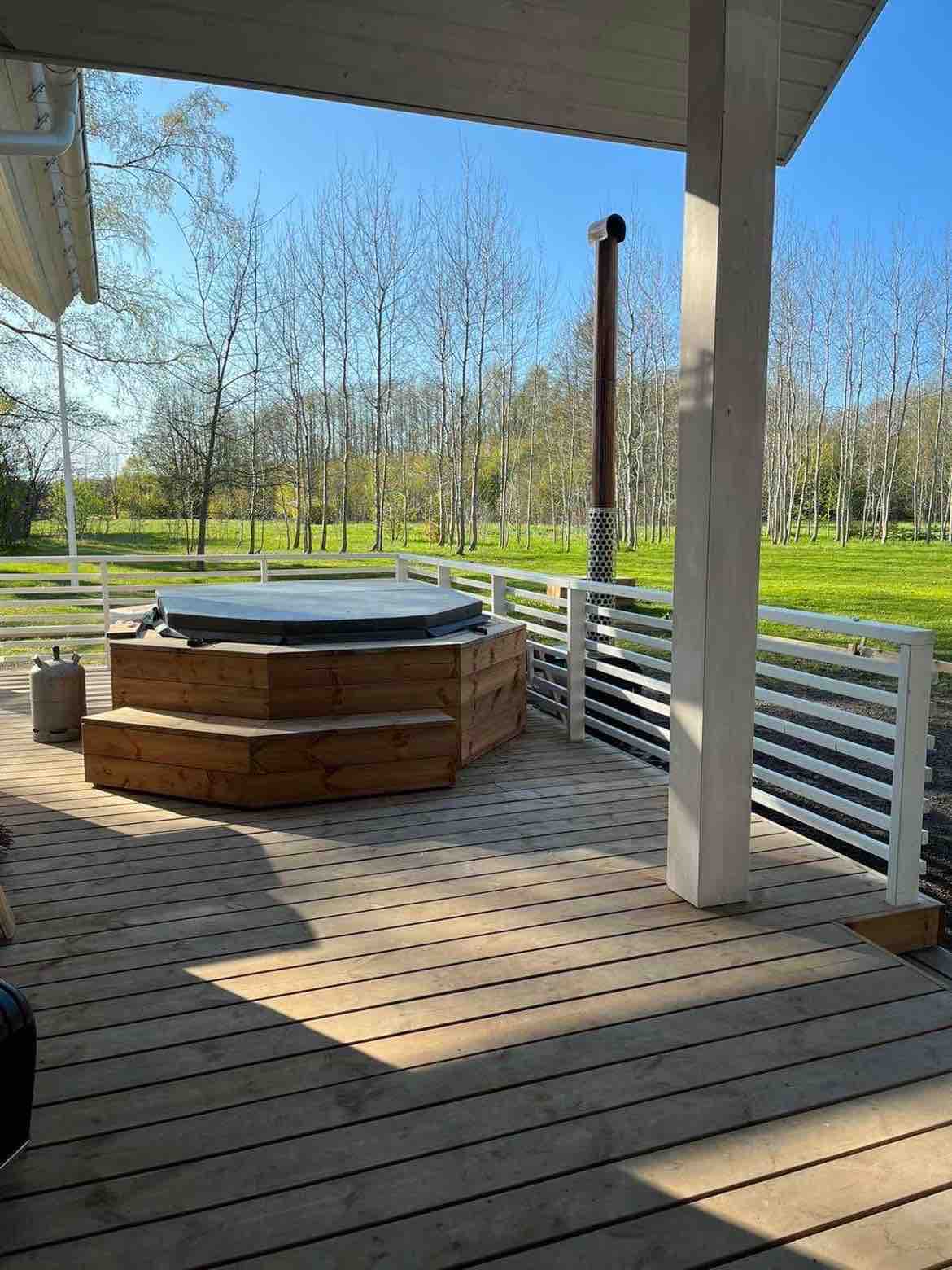 A wooden deck features a hot tub surrounded by natural scenery. Sunlight filters through nearby trees, illuminating the spacious outdoor area. The deck is enclosed by white railing, enhancing the open feel.
