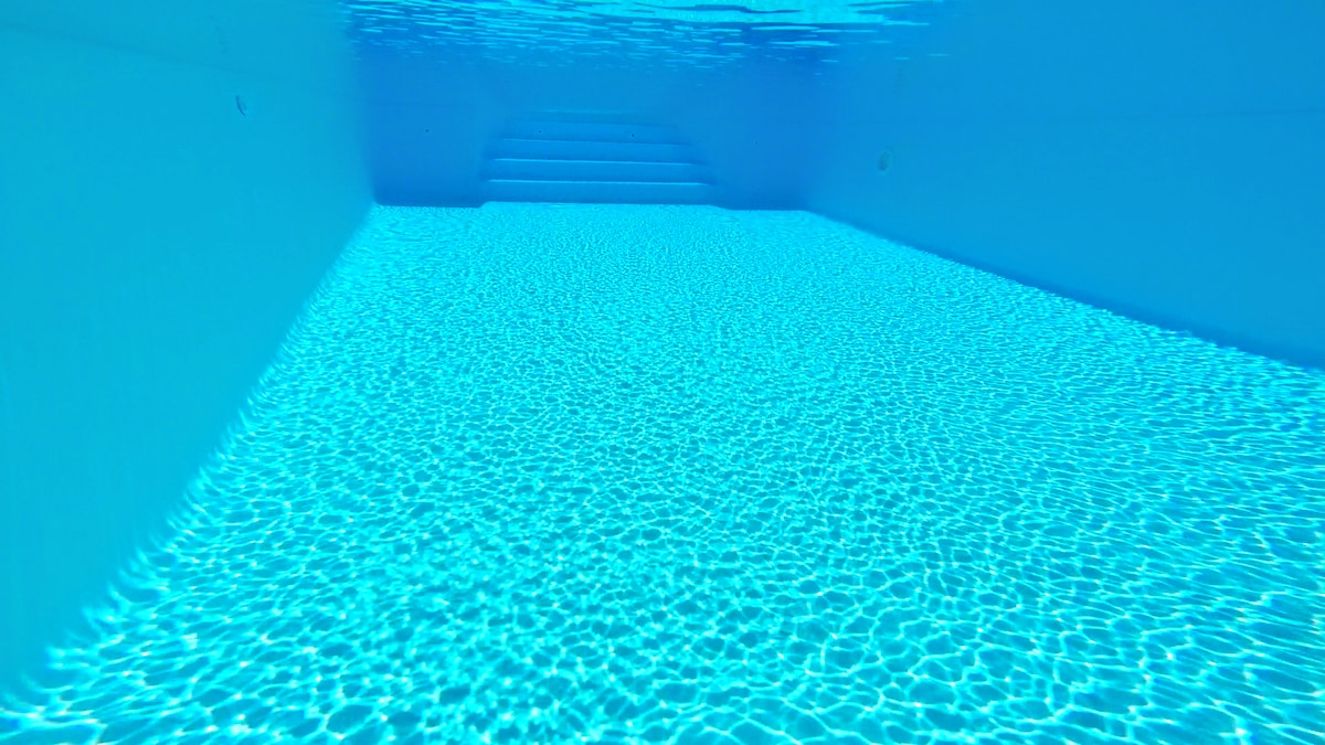 The crystal-clear swimming pool features gently rippling water reflecting light above. Steps lead down into the pool, providing easy access. The overall atmosphere suggests a refreshing and calm environment for relaxation.