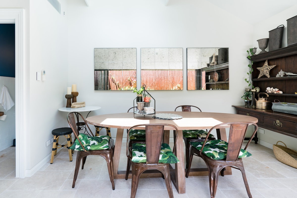 A bright dining area features a large wooden table surrounded by several dark wooden chairs with green tropical-patterned cushions. Mirrors on the wall reflect the room's light. A rustic sideboard displays decorative items, adding to the space's charm.