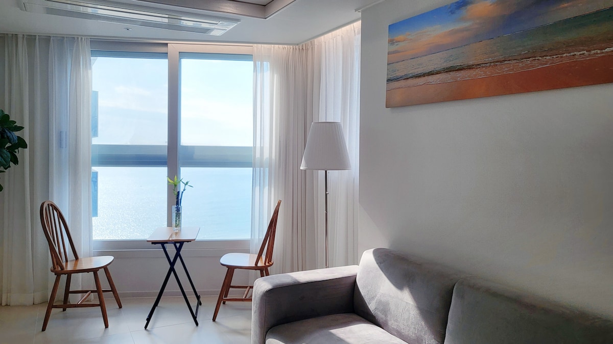 A bright living area is highlighted by large windows offering a sea view. A small table is set for two, accompanied by two wooden chairs. A sofa provides comfortable seating, and a floor lamp adds soft lighting. The walls are adorned with a serene coastal art piece.