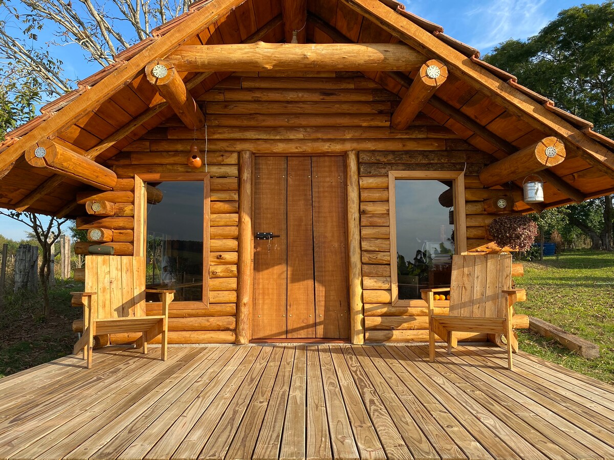 A wooden cabana with a charming log exterior is displayed. Two wooden chairs are positioned on the spacious deck, offering a place to relax. Large windows on either side of the entrance provide views of the surrounding nature.
