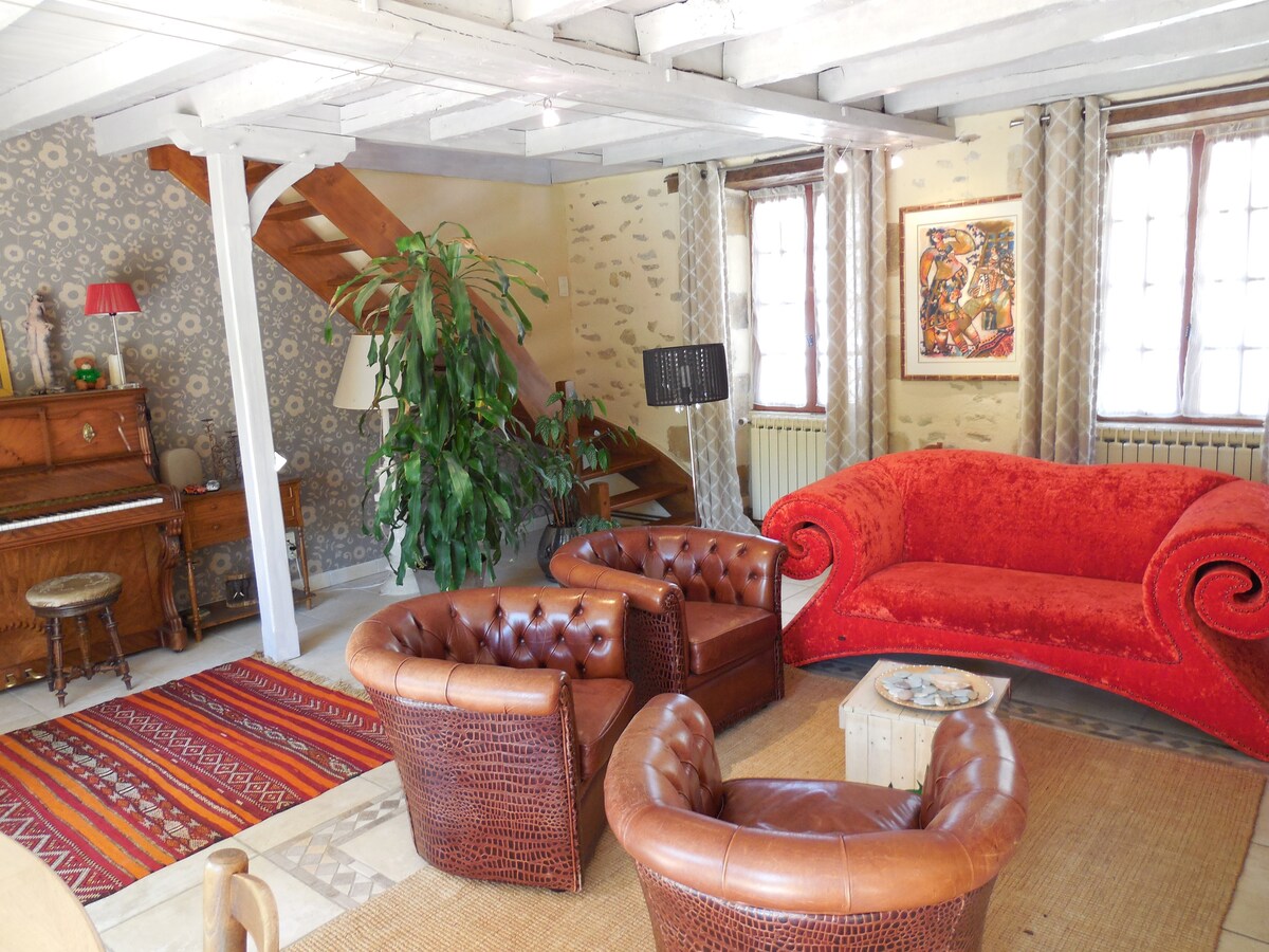 The living area features elegant design elements, including a vibrant red sofa and two armchairs with intricate woven patterns. A piano and a plant enhance the inviting atmosphere. Light enters through windows dressed with patterned curtains, while a colorful rug adds warmth to the space.