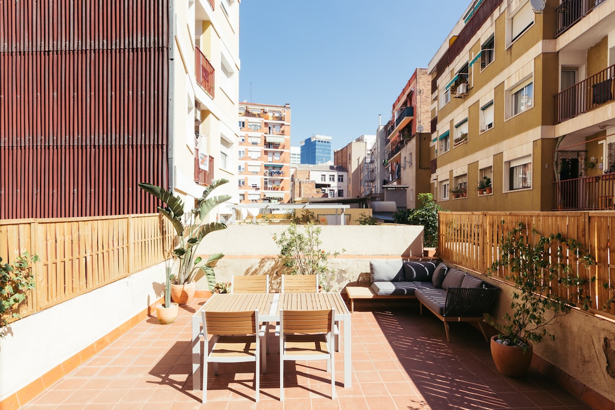 A rooftop terrace overlooks neighboring buildings, featuring a mix of potted plants and outdoor furniture. A wooden table and chairs are positioned beside a comfortable sofa, while terracotta tiles cover the area. Sunlight brightens the space, creating an inviting outdoor setting.