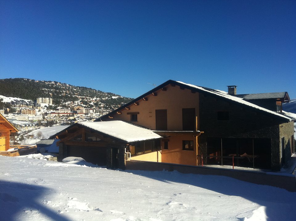 A snow-covered chalet is set against a clear blue sky, with a sloped roof and several windows. Nearby, the rolling snow-covered hills and distant village buildings create a serene mountain landscape.