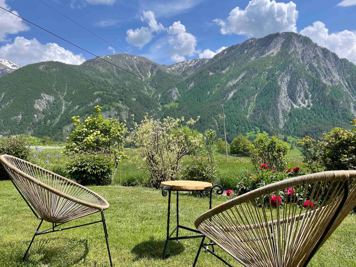 Two woven chairs are positioned on a grassy area, offering a view of rolling mountains and a vibrant landscape. A small table sits between the chairs. Lush greenery and blooming flowers are visible, enhancing the serene outdoor setting.