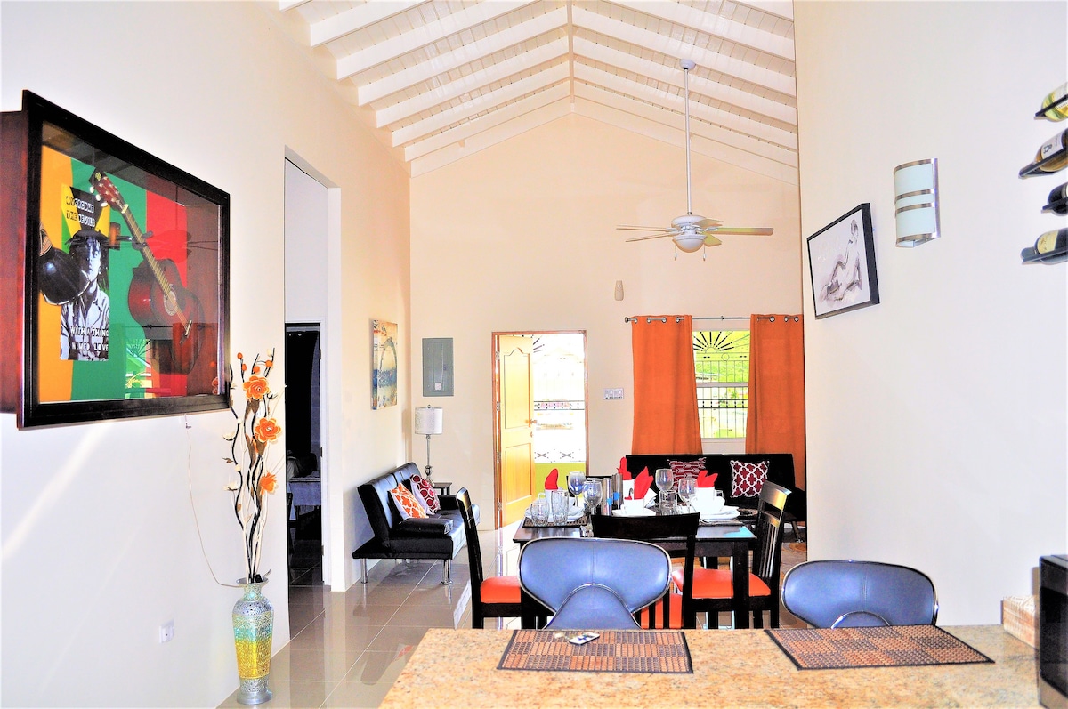 A spacious living and dining area is highlighted by high ceilings and an open layout. A dining table is set for multiple guests, surrounded by colorful accents. A sofa and side tables provide a welcoming seating space, enhancing the atmosphere of relaxation and comfort.