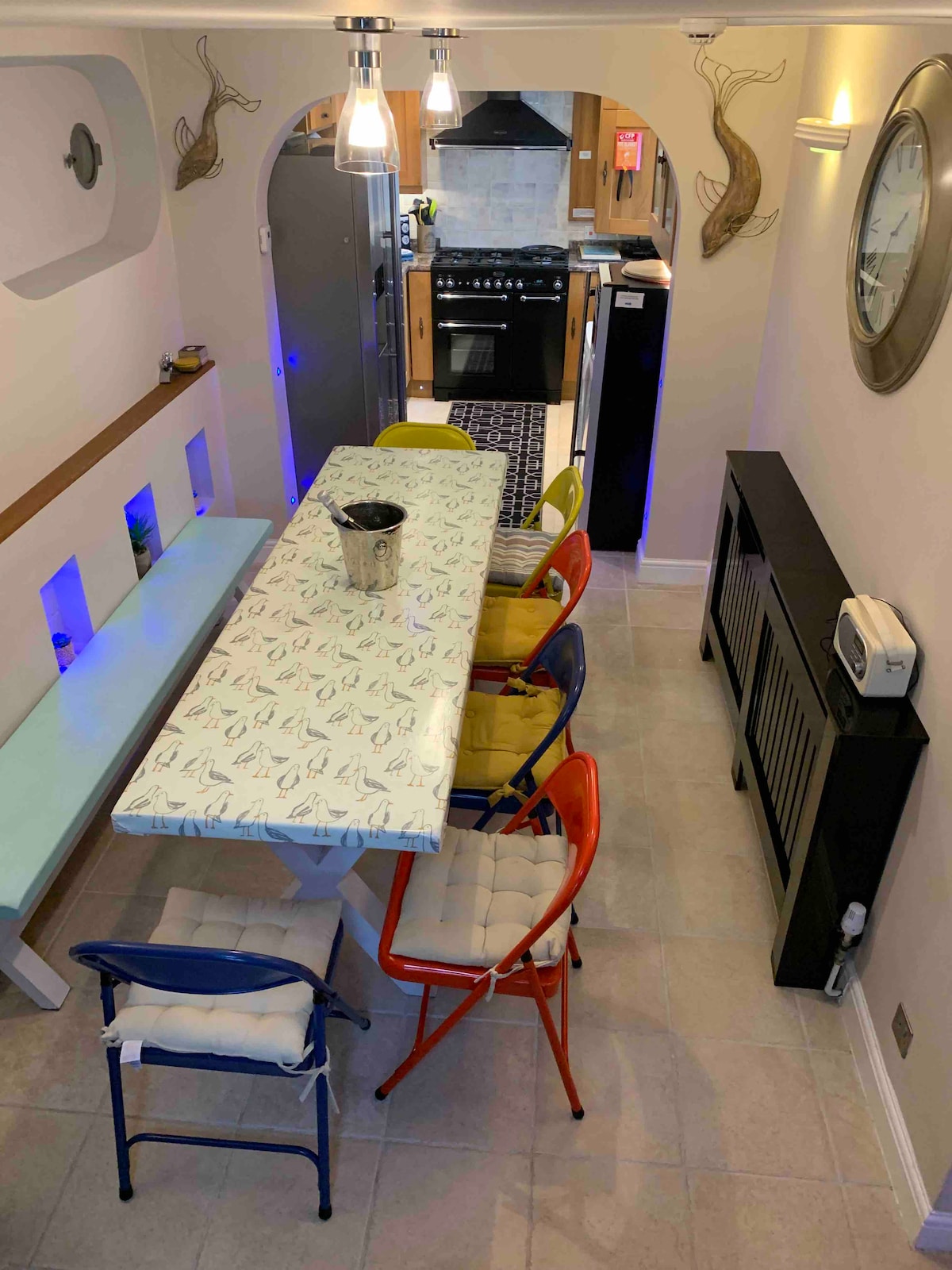 A modern dining area features a long table with a playful fish-patterned top, surrounded by colorful chairs in blue, yellow, red, and beige. Soft lighting is provided by hanging pendant fixtures, and the space includes a stylish radiator and wall decor.