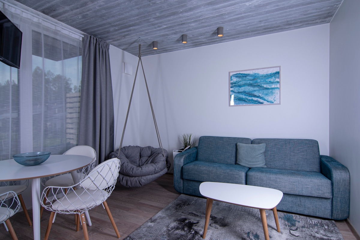 A modern living area features a comfortable blue sofa and a unique indoor swing. A round dining table is accompanied by white chairs. Natural light enters through large curtains, and calming artwork is displayed on the wall, enhancing the serene environment.