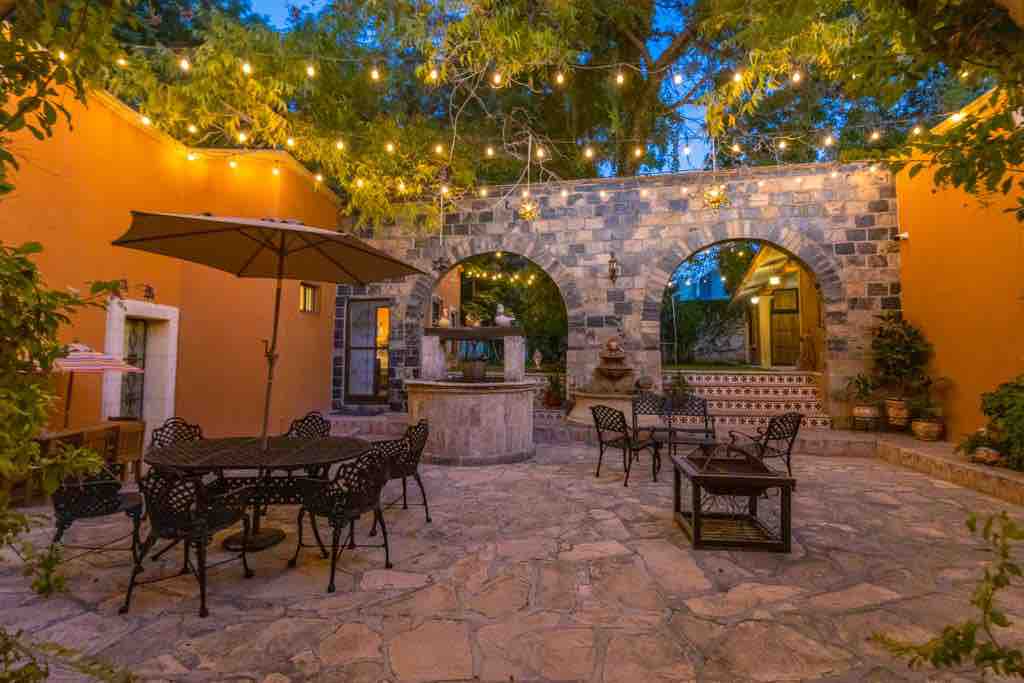 A charming outdoor patio area is featured, surrounded by vibrant orange walls and illuminated by string lights. Stone arches create an inviting focal point, while metal tables and chairs provide seating. A small fountain adds a serene touch to the space, enhancing its social atmosphere.