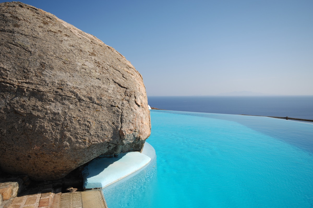 An infinity swimming pool extends towards the horizon, seamlessly blending with the azure sea. A large natural rock formation is positioned at the pool's edge, enhancing the tranquil ambiance. The clear sky complements the serene blue water, offering a picturesque view of the surrounding landscape.