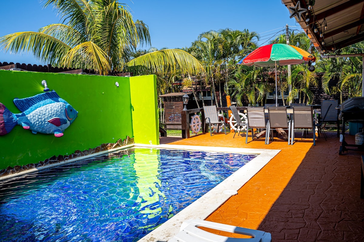 A bright pool area is framed by lush palm trees and colorful decor. The swimming pool features sparkling blue water, while a vibrant green wall adds a playful touch. A set of outdoor furniture and a large umbrella are positioned nearby for relaxation.
