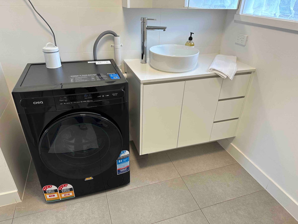 A modern laundry area is displayed, featuring a black washing machine with control panel on the left. To the right, a sleek white vanity with a circular basin is positioned above three drawers, complemented by a clean towel neatly placed beside the basin.
