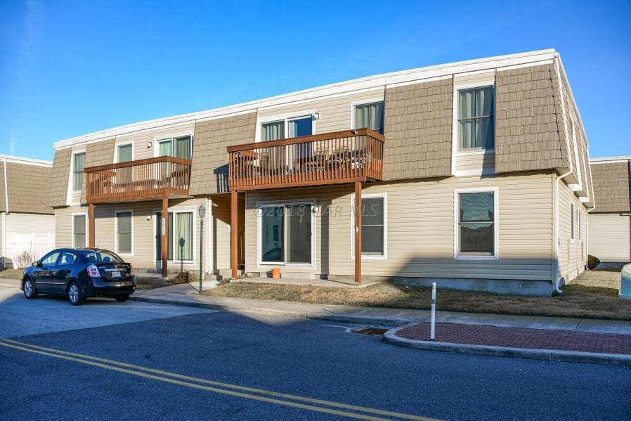 The two-story building features a light exterior with a shingled roof. Two balconies are present on the upper level, overlooking a paved area. Parking spaces are visible in front, along with a wide street providing access to the property.
