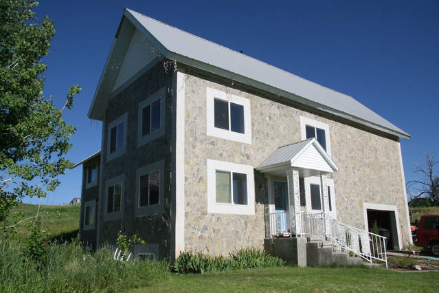 The exterior of a two-story stone house is shown, featuring multiple large windows that invite natural light. A white front entryway with steps leads to the main entrance, surrounded by a well-maintained lawn and greenery. The skyscape is clear and blue.
