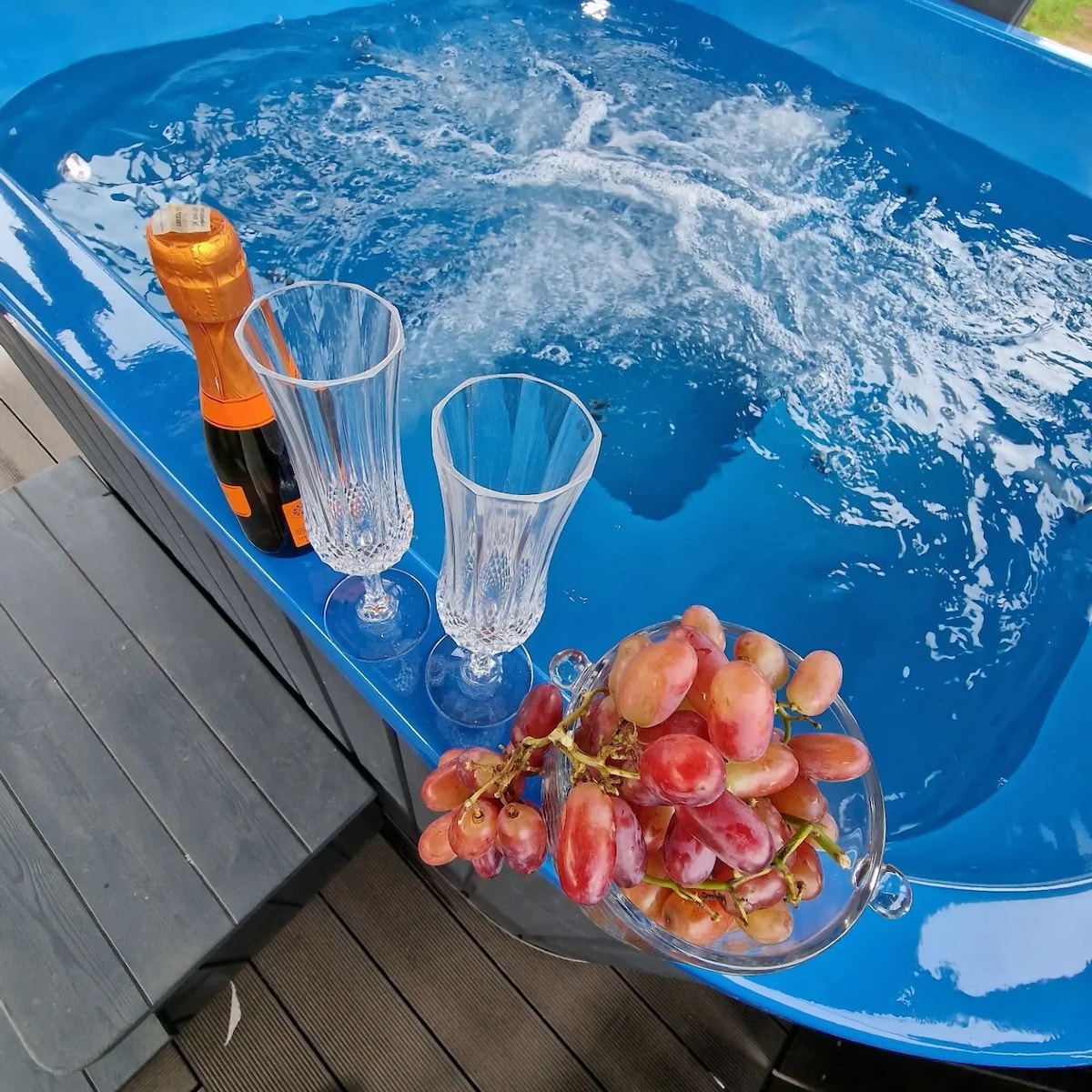 The image features a blue hot tub with bubbles rising to the surface. Nearby, two elegant glass flutes sit next to an open bottle of sparkling wine. A glass dish filled with grapes adds a touch of freshness, all set against a backdrop of wooden decking.
