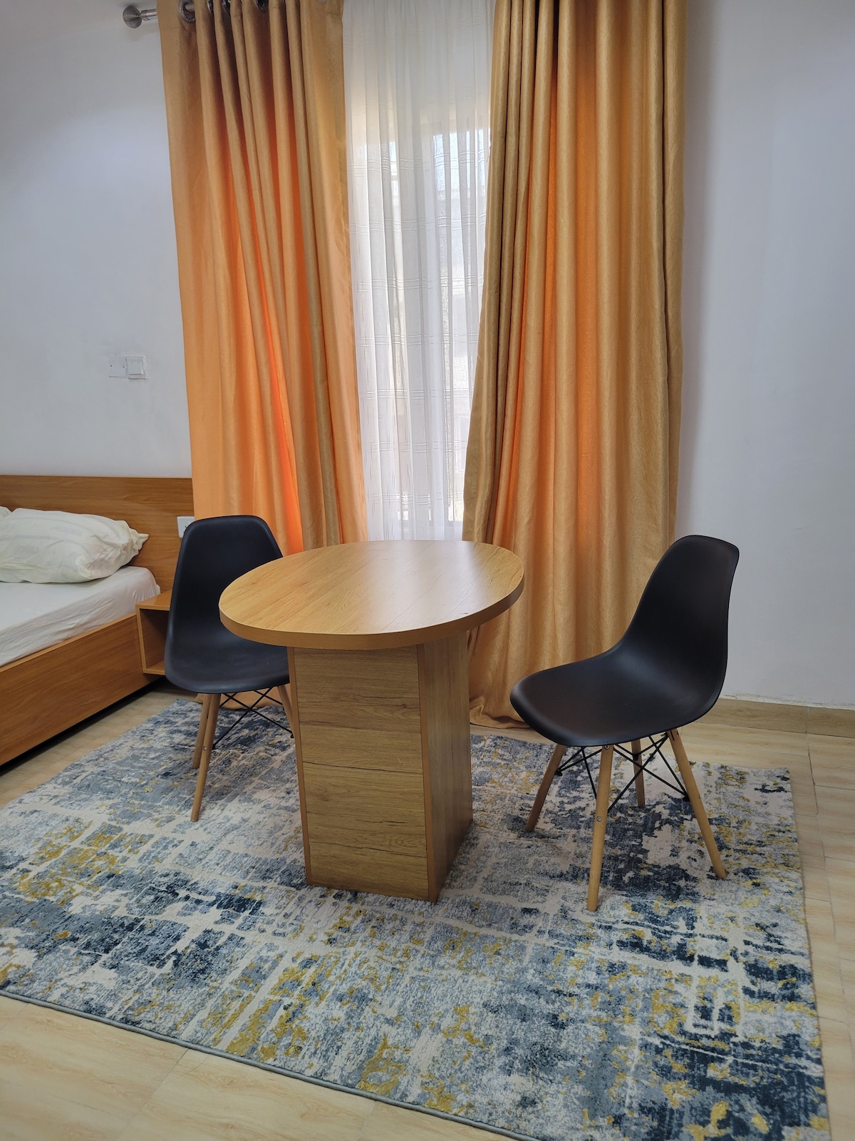 A small dining area is featured with a round wooden table and two black chairs. Soft orange curtains frame a window, allowing natural light to brighten the space. A patterned rug adds warmth to the flooring, complementing the neutral tones of the room.