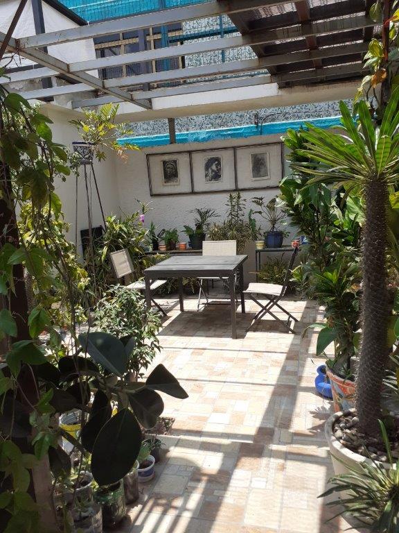 An outdoor patio area is surrounded by various potted plants and leafy greenery. A small table with two chairs provides a seating option, while shaded structures offer partial cover. Decorative elements like wall art enhance the serene environment.
