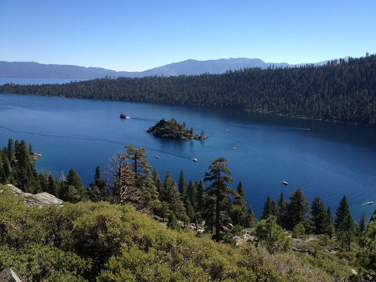 A view of Lake Tahoe showcases its calm, blue waters surrounded by a forested shoreline. A small island is visible in the center of the lake, with boats gently floating nearby. The landscape features tall pine trees in the foreground, providing a natural frame for the scenic backdrop.