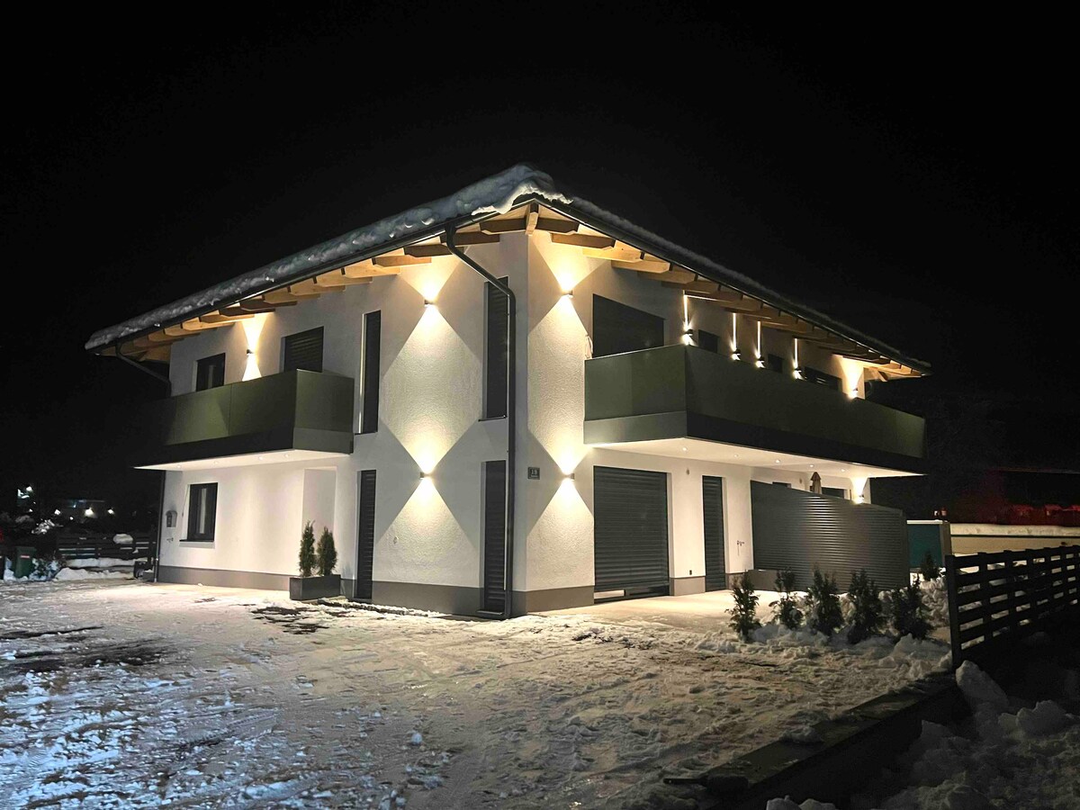 A contemporary building is illuminated by exterior lights, highlighting its architectural features. Snow blankets the ground, and the structure includes balconies on the upper level. The entrance is clear, and minimal landscaping is visible along the exterior.