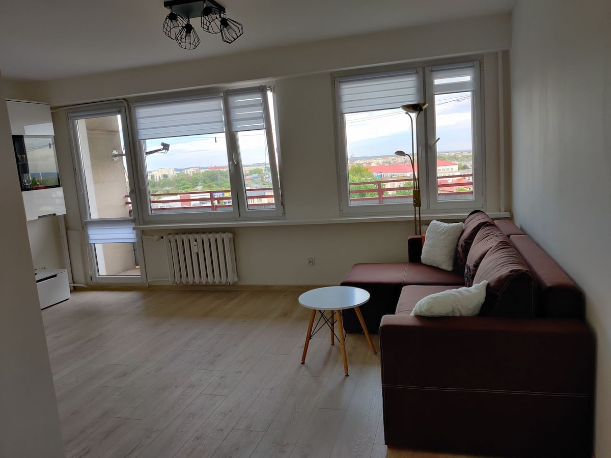 A bright living area features a spacious sofa with cushions, a small round coffee table, and large windows allowing natural light. A sleek lamp stands beside the sofa, and a partial view of the surrounding landscape is visible through the window.