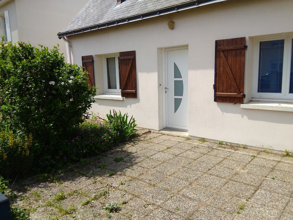The exterior of the house is shown, featuring a well-maintained front garden with green shrubs and blooming flowers. The entrance includes a white door with a decorative window design, framed by wooden shutters. A stone pathway leads to the entrance, providing a clear and welcoming entryway.