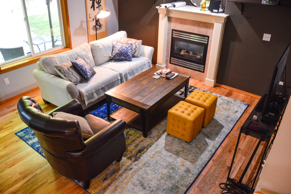 A cozy living area features a comfortable light gray sofa accented with decorative pillows. A rustic coffee table sits on a large area rug, accompanied by two yellow poufs. A sleek fireplace serves as a focal point, and a television is positioned nearby.