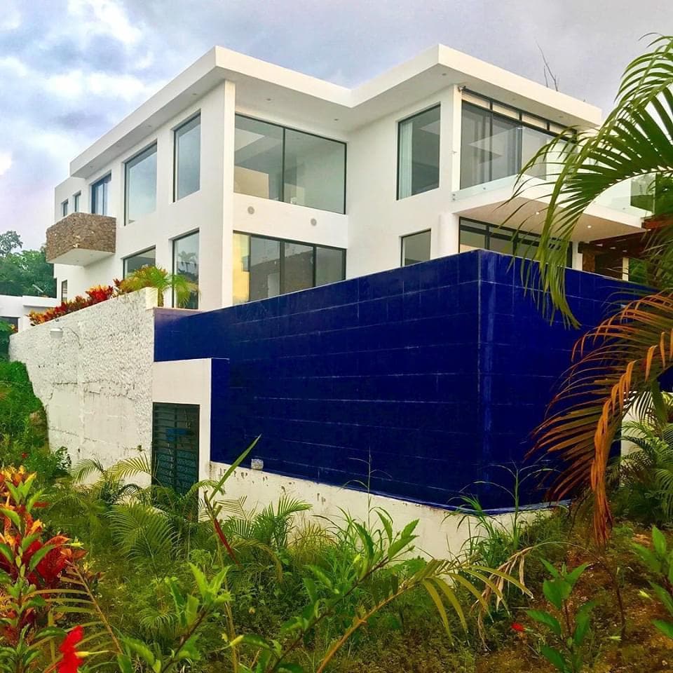 A modern two-story house is framed by lush green landscaping, featuring large glass windows that brighten the exterior. The building’s façade is complemented by a striking blue wall, creating a bold contrast with the surrounding nature.