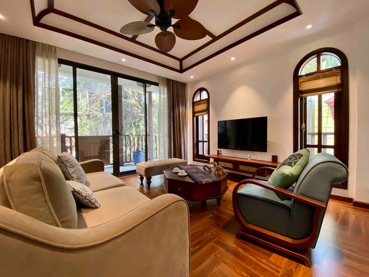 The living room features large windows allowing natural light to fill the space. Comfortable seating is provided by a light-colored sofa and an armchair. A coffee table is positioned centrally, with a flat-screen television mounted on the wall, offering ample space for relaxation.