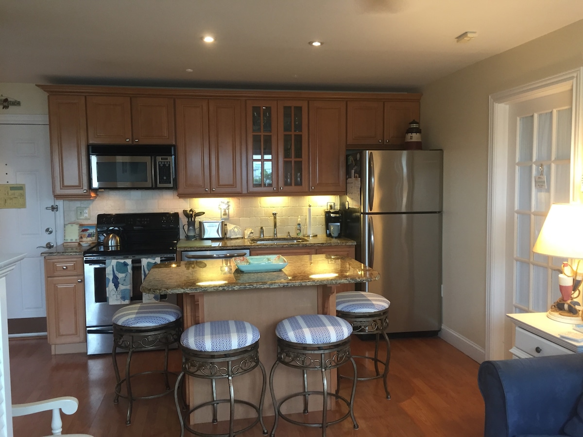 A modern kitchen features wooden cabinetry and granite countertops, equipped with stainless steel appliances including a refrigerator, electric range, and microwave. Three stools with blue-and-white upholstery are positioned at the island, with under-cabinet lighting highlighting the workspace.
