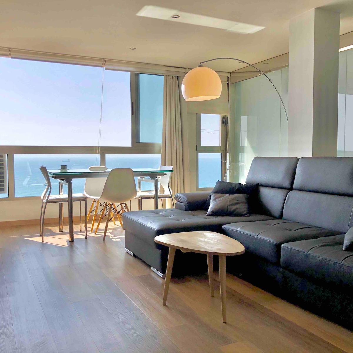 A modern living space is featured with a black sectional sofa and a small round coffee table. A dining area with a glass table and white chairs offers views of the sea through large windows, allowing natural light to fill the room.