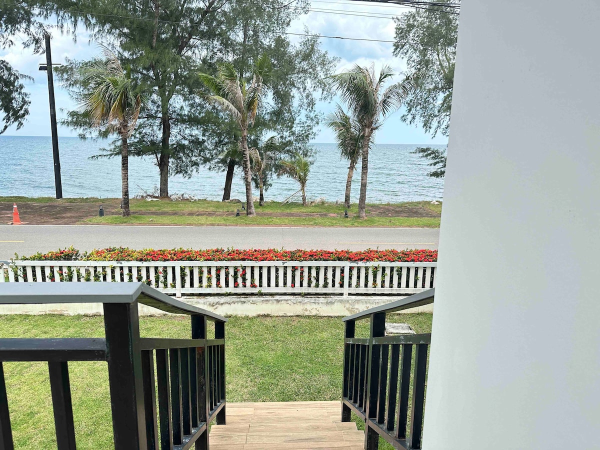 The view from a set of stairs includes a well-maintained lawn leading to a vibrant flowerbed. Palm trees sway gently in the breeze, while the ocean is visible beyond the road, inviting a sense of calm and relaxation.