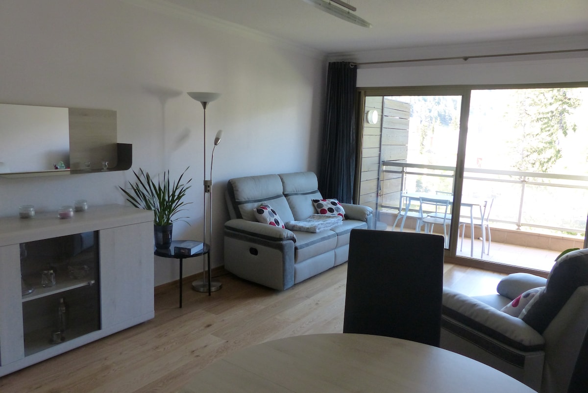 A bright living area features a comfortable gray sofa, accent chairs, and a dining table. A large glass door opens to a balcony with outdoor furniture. Natural light enhances the space, complemented by minimal decor and a potted plant.