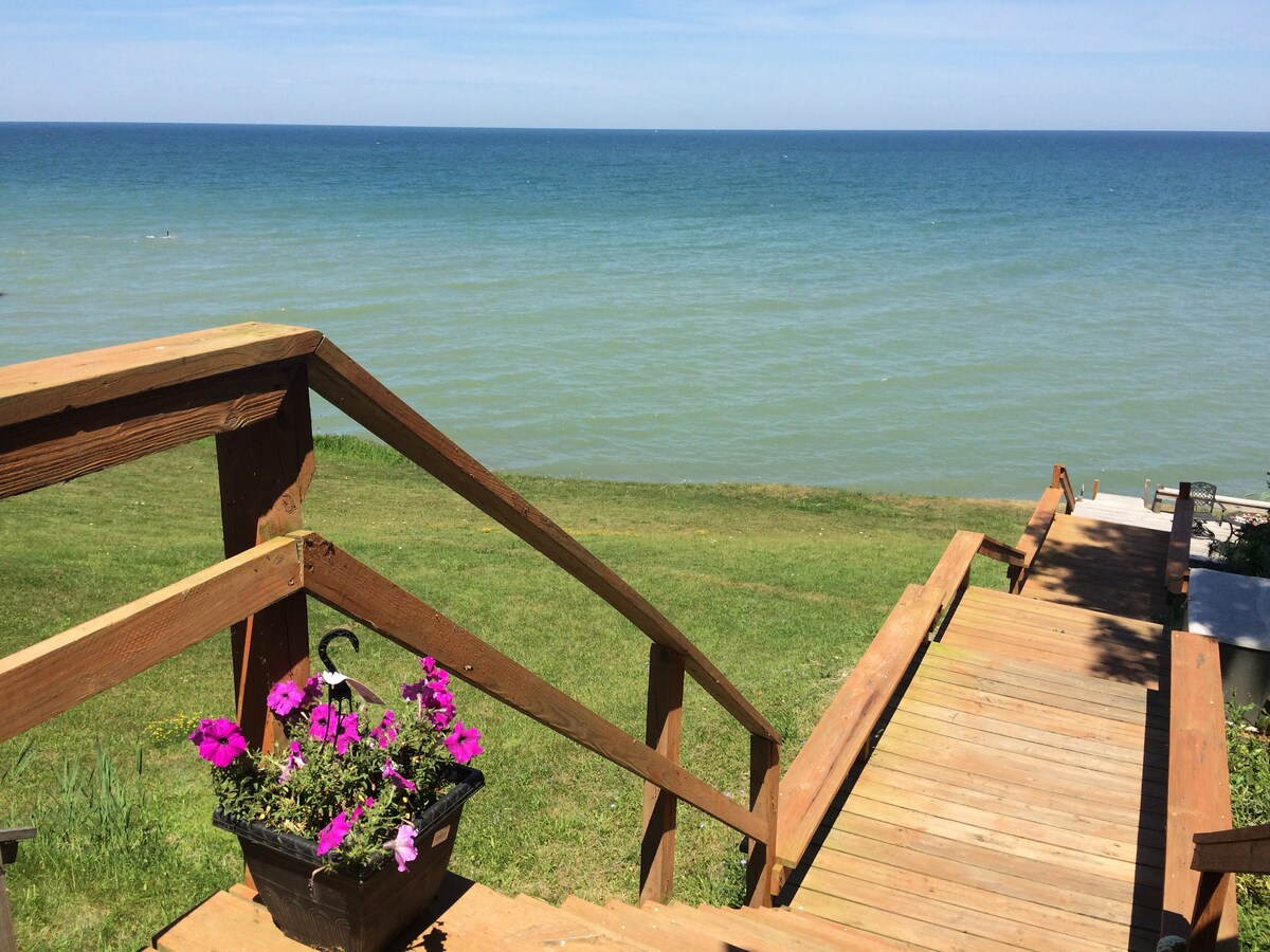 A wooden staircase leads down from the deck to a grassy area, offering a clear view of Lake Erie. Vibrant pink flowers are situated in a hanging basket, complementing the serene blue waters and distant horizon.