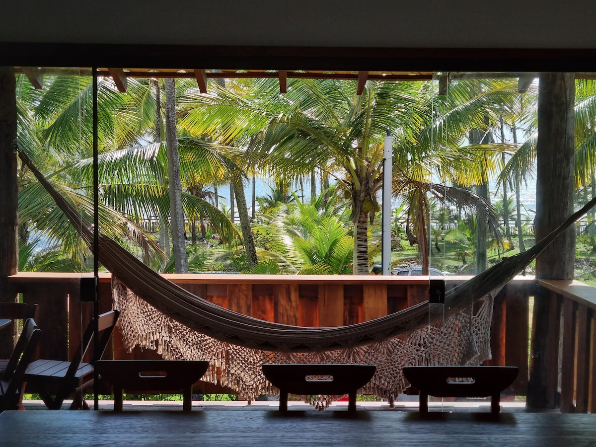 A comfortable hammock is suspended on a wooden balcony, surrounded by lush palm trees. The ocean can be glimpsed through the dense foliage, providing an inviting view. Two wooden benches are positioned nearby, enhancing the inviting outdoor space.
