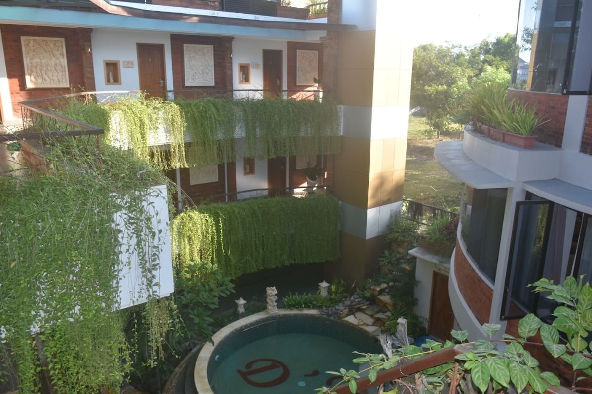 A serene courtyard is surrounded by lush green vines draping over balconies. A circular water feature, accented by sculptural elements, is nestled among the greenery, providing a calming focal point within the space. Sunlight filters through, illuminating the vibrant outdoor setting.