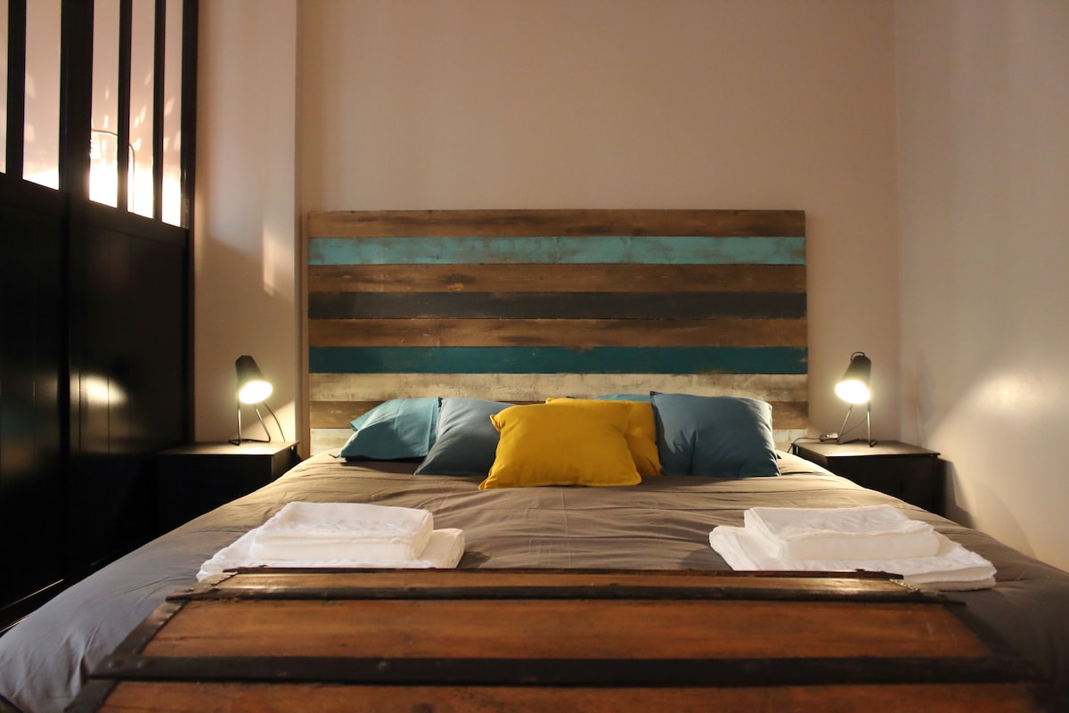 A spacious bed is showcased with a wooden headboard featuring a striped design in natural tones and turquoise. Soft blue and yellow pillows complement the neutral bedding. A pair of bedside lamps is positioned on either side, illuminating the inviting sleeping area.