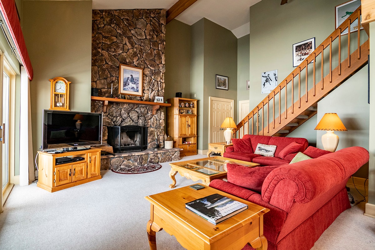 A spacious living area features a large stone fireplace, surrounded by wooden furniture. A red sofa and matching armchair provide seating, while a wooden coffee table sits in front. Natural light enters through large windows, illuminating the carpeted floor and decorative items.