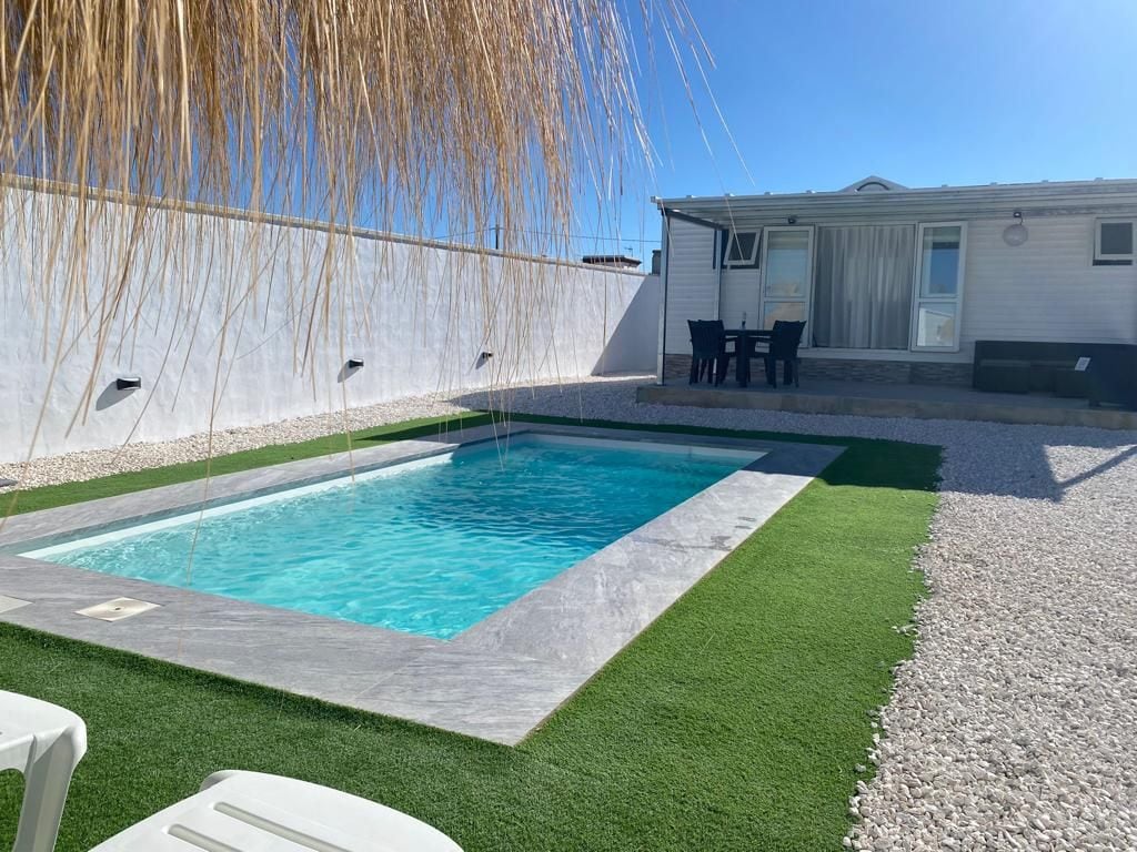 A private garden area displays a sparkling pool surrounded by manicured grass. Sun loungers are positioned nearby for relaxation. Behind the pool, a porch houses a dining table and chairs, creating an inviting outdoor space under clear blue skies.