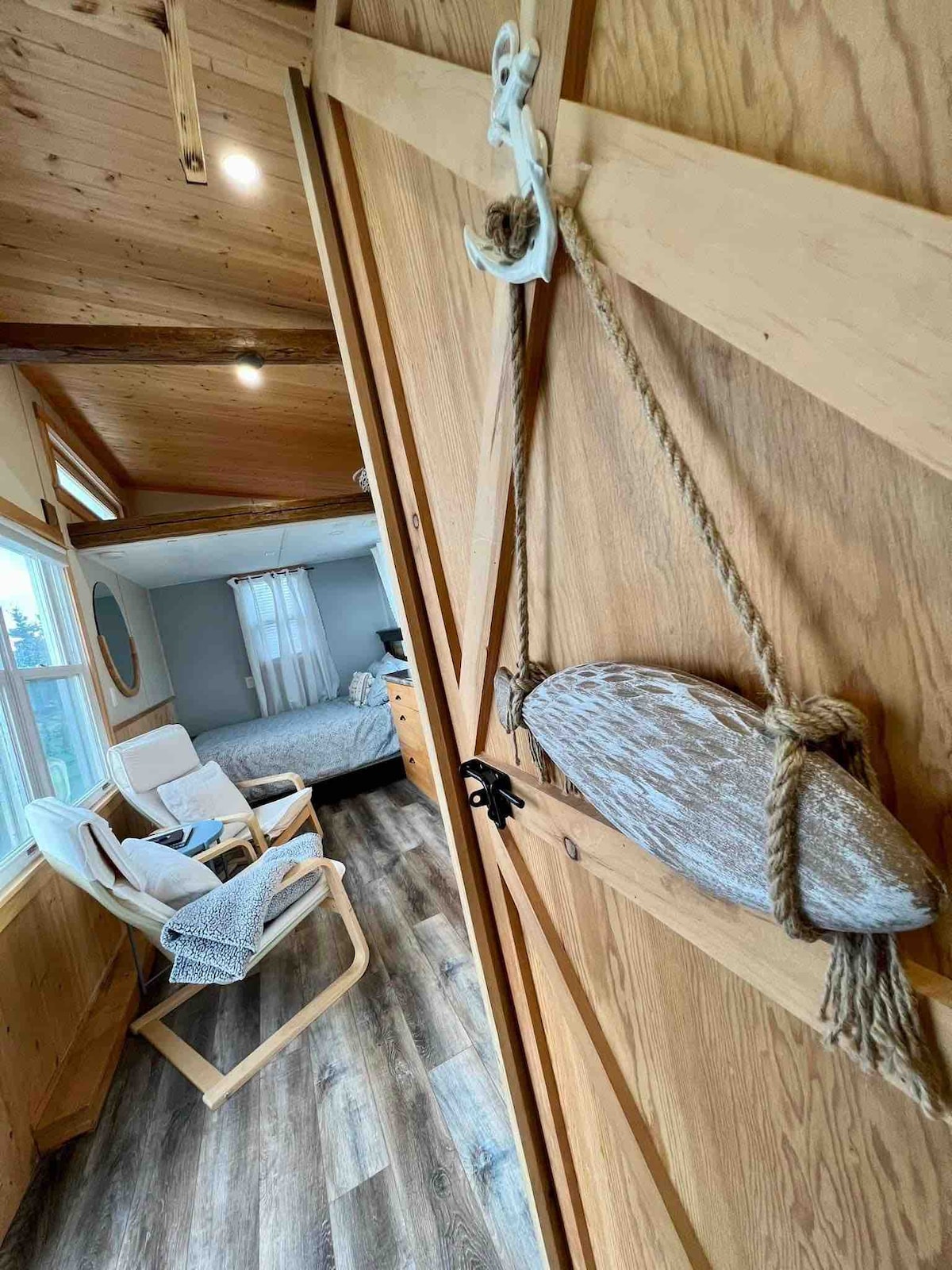 A wooden sliding door is featured at the entrance, accented by a decorative piece resembling a seashell hung from rope. In the background, a cozy bedroom is visible, complete with a spacious bed and natural light filtering through a window.