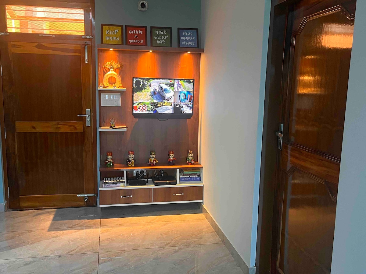 An entryway is presented with modern decor, featuring a mounted television above a wooden console. A combination of decorative items, including figurines, adds character. A warm light spills from the shelf, enhancing the ambiance while a sturdy door is visible at the far end.