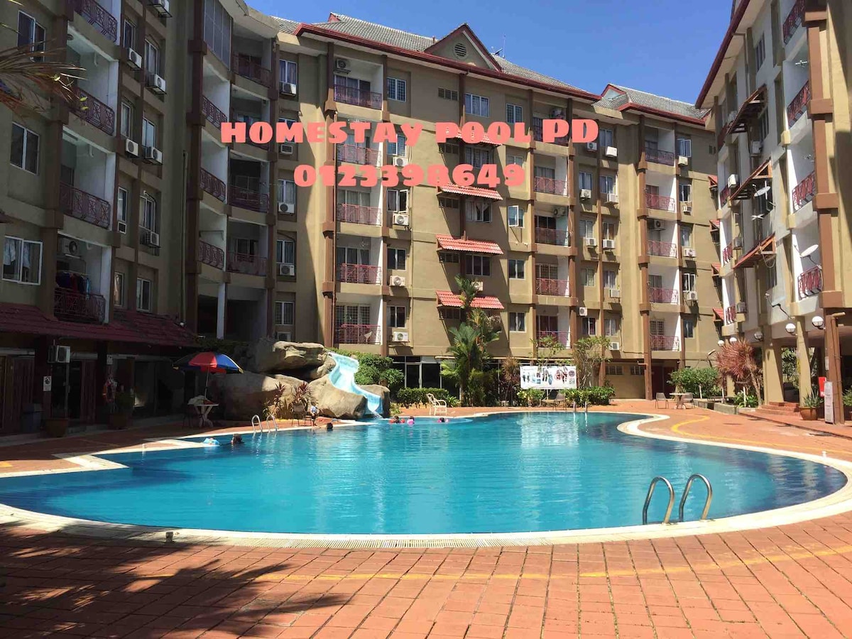 The image features a large swimming pool surrounded by a spacious paved area. The pool is rectangular with clear blue water. Apartment balconies with plants are visible in the background, and some guests can be seen enjoying the area under the sun.